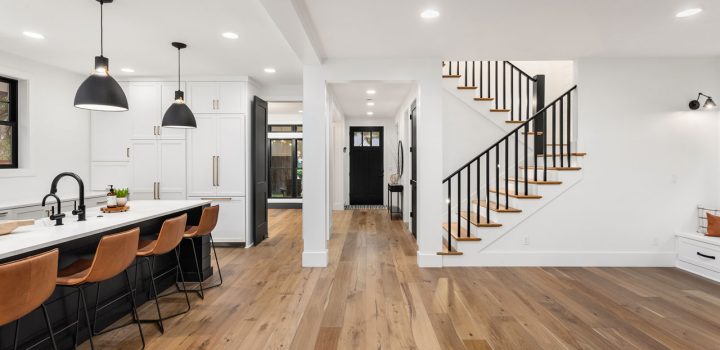 Contemporary open-concept kitchen and entryway featuring wide-plank light oak hardwood, white cabinetry, black island and pendant lights, and a sleek black-and-wood staircase—showcasing seamless flow and design cohesion.