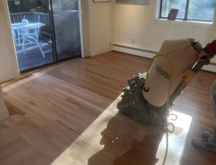 Technician operating a Lägler drum sander on light oak hardwood flooring in a sunlit room with sliding glass doors; yellow hose and dust collection bag visible—showcasing professional, low-dust refinishing.