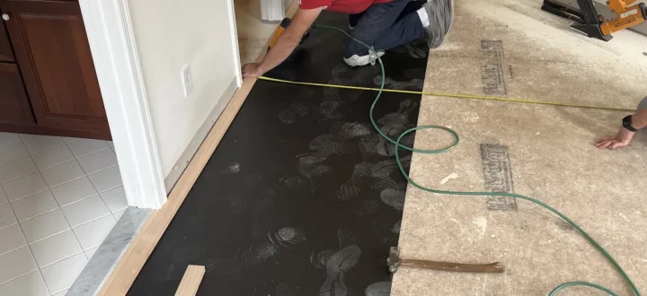 Installer in red shirt kneeling to fit hardwood planks along a transition strip near a tiled bathroom; black underlayment, pneumatic nailer, utility knife, and green air hose visible—highlighting attention to detail at thresholds.