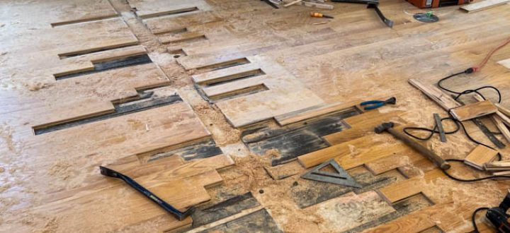 Mid-renovation kitchen scene showing partially removed hardwood planks exposing subfloor; tools, debris, and exposed wall framing indicate active floor replacement; cabinetry and appliances remain in place.