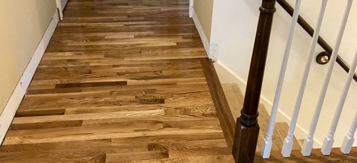 A clean, polished medium-tone oak hardwood hallway leading toward a white door, bordered by cream walls and white baseboards; dark wood staircase railing with white spindles visible on the right.