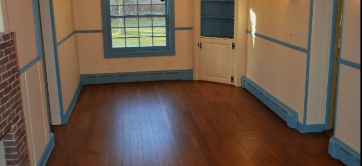 Deep chestnut-finished hardwood floor in a traditionally styled room with cream walls, blue-painted trim, built-in shelving, a brick hearth, and a vintage chandelier.
