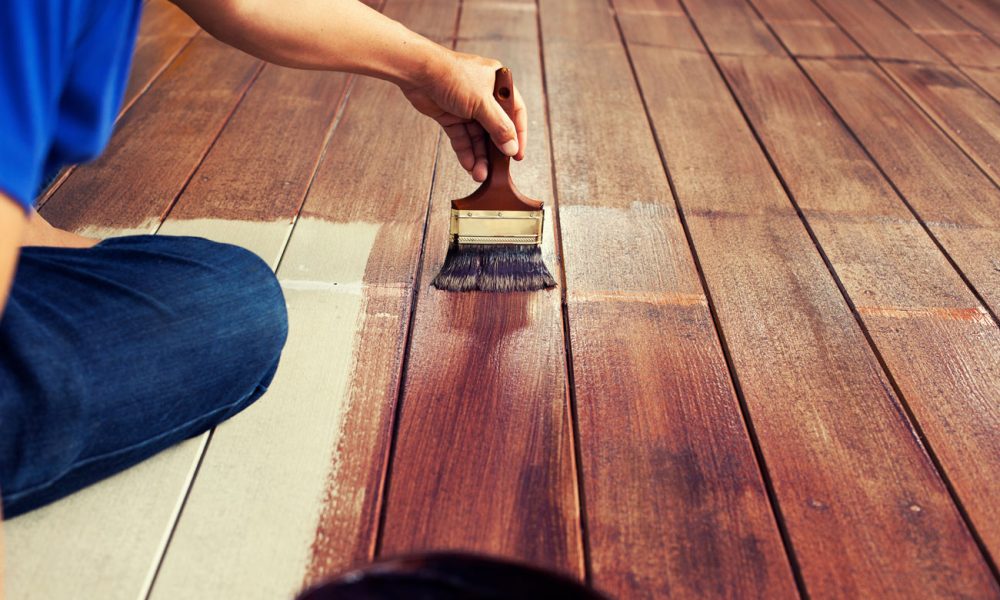 Close of a worker applying rich brown stain with a natural-bristle brush onto sanded hardwood floorboards; visible contrast between treated and untreated sections shows transformation in progress.