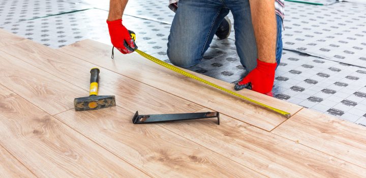 Installer in red gloves measuring and fitting light wood-grain LVP planks over patterned underlayment; tools include tapping block, hammer, and tape measure—demonstrating proper floating-floor technique.