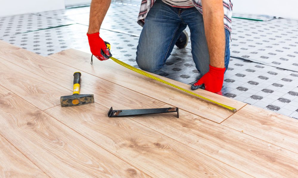 Installer in red gloves measuring and fitting light wood-grain LVP planks over patterned underlayment; tools include tapping block, hammer, and tape measure—demonstrating proper floating-floor technique.
